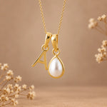 Gold initial pendant necklace featuring the letter 'A' and a teardrop-shaped white pearl, shown against a warm beige background with dried flowers.