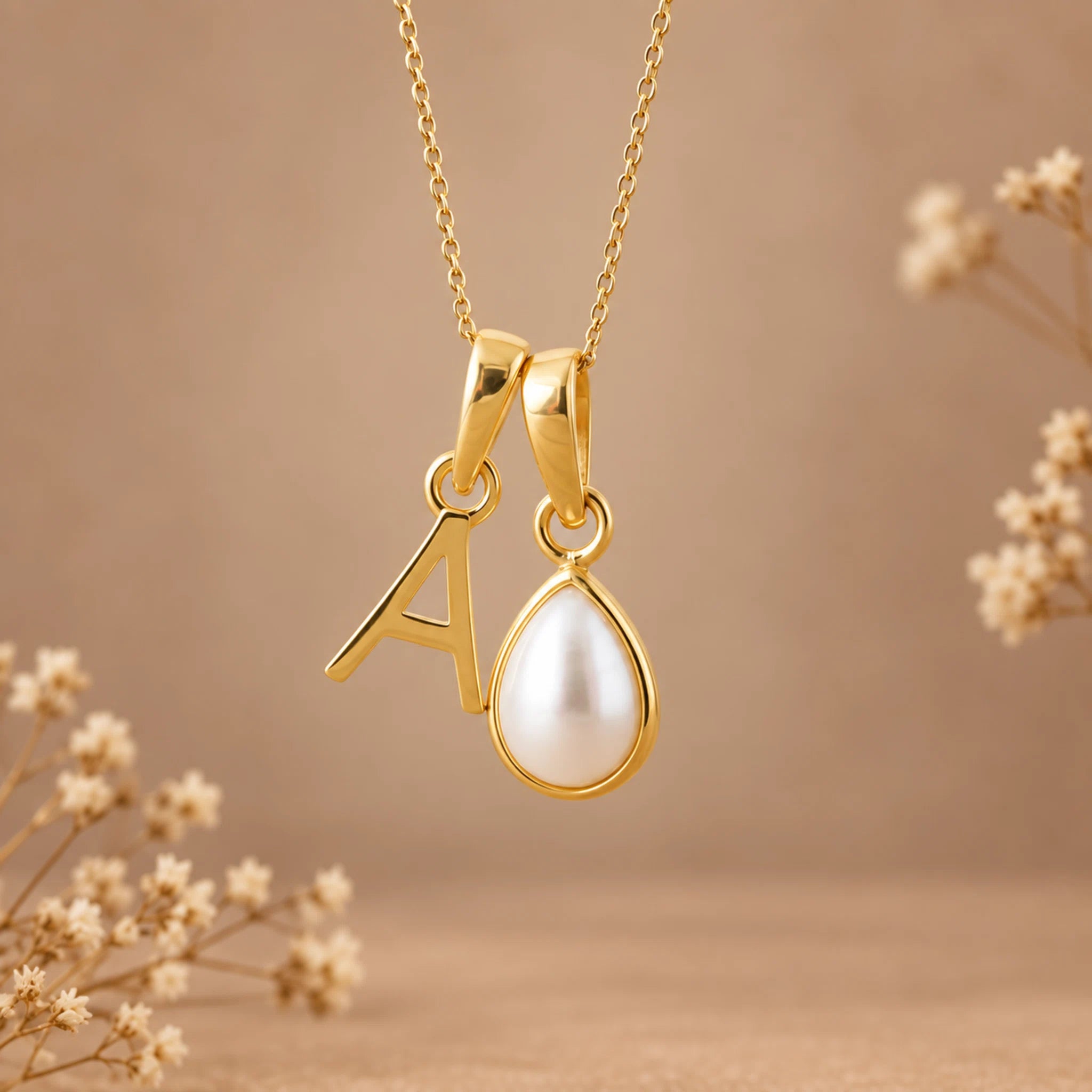 Gold initial pendant necklace featuring the letter 'A' and a teardrop-shaped white pearl, shown against a warm beige background with dried flowers.