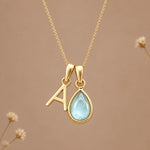 A gold initial necklace featuring a letter 'A' pendant and a teardrop-shaped light blue gemstone pendant on a delicate gold chain, set against a warm beige background with subtle dried flowers.