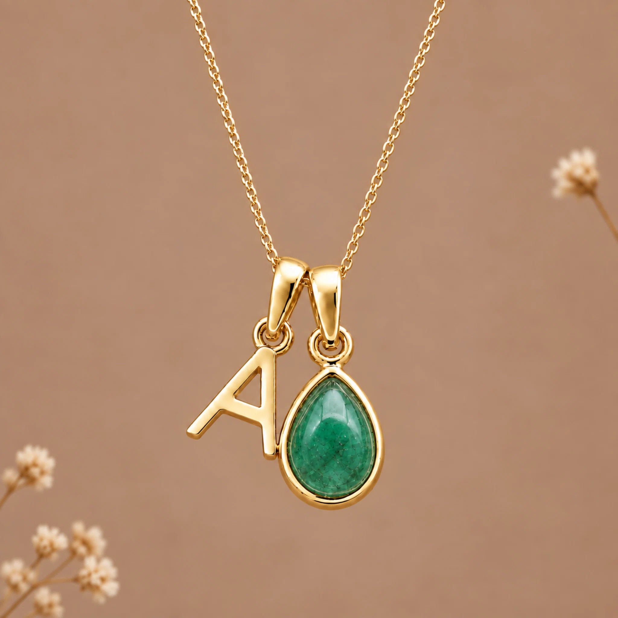 Gold initial necklace with a teardrop-shaped green emerald stone pendant and a gold letter 'A' charm, shown on a warm brown background with small dried flowers.