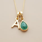 Gold necklace with 'A' pendant and green emerald teardrop pendant on a beige background
