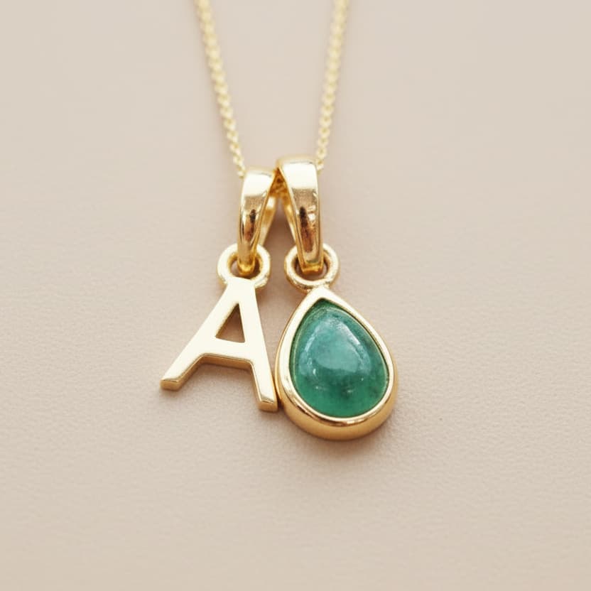 Gold necklace with 'A' pendant and green emerald teardrop pendant on a beige background