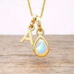 Gold necklace with a teardrop-shaped pendant and letter 'A' charm on a wooden surface.