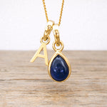 Gold necklace with a blue sapphire teardrop pendant and letter 'A' charm on a wooden surface.