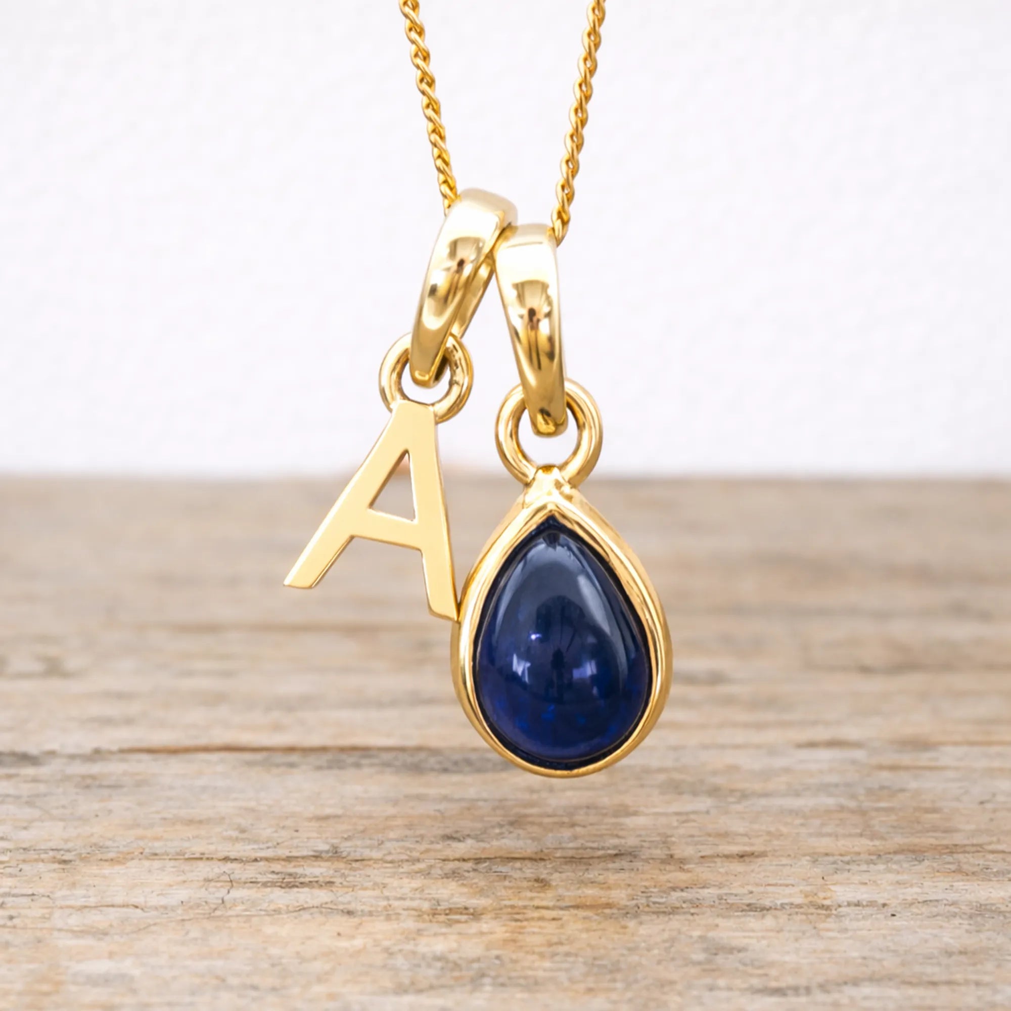 Gold necklace with a blue sapphire teardrop pendant and letter 'A' charm on a wooden surface.
