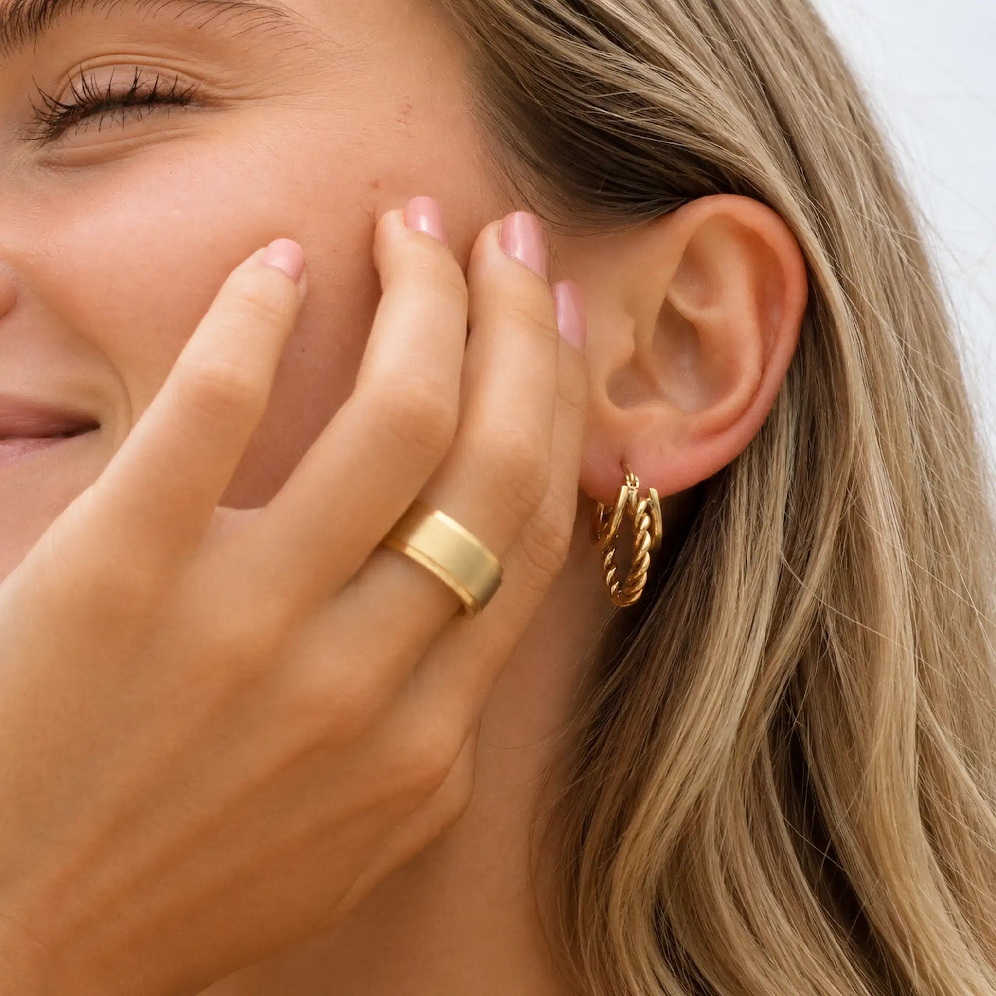 Close-up of a woman wearing a thick gold band ring on her finger and small gold hoop earrings in her ear, with her hand resting near her face.