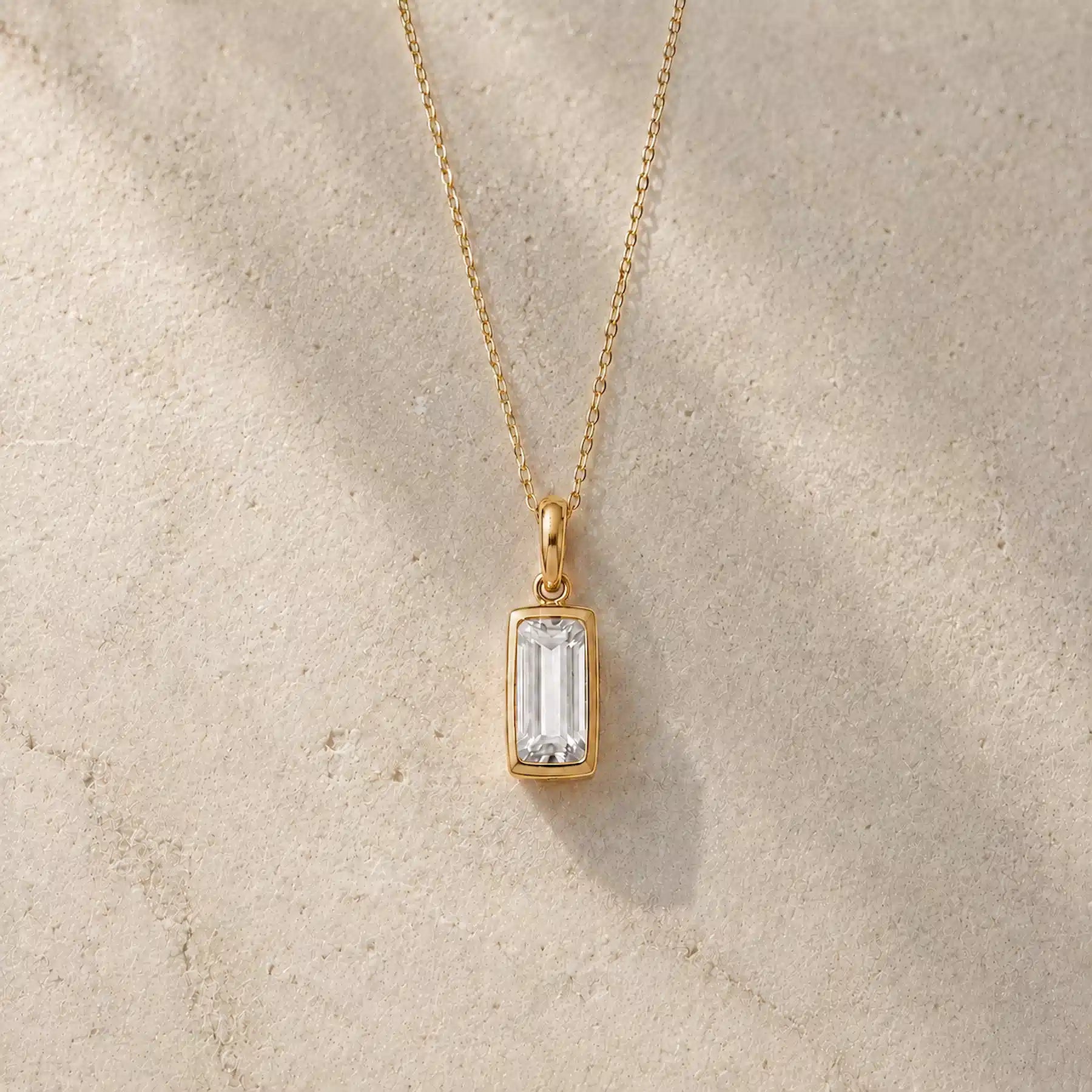 Gold fine chain necklace with a rectangle faceted bezel set april cubic zirconia pendant on a light stone background.