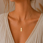 aquarius necklace - A close-up of a woman's neck and chest wearing a delicate gold chain necklace with a small rectangular gold pendant featuring a minimalist engraved aquarius star sign line glyph symbol, styled with a cream-colored lace-trimmed top.