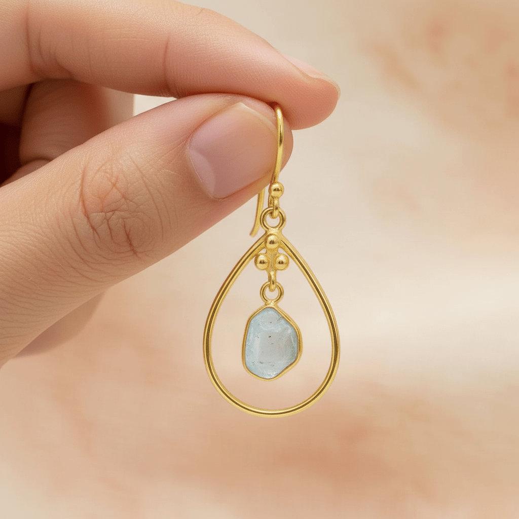Gold earring with a natural raw aquamarine gemstone held by a hand against a blurred background