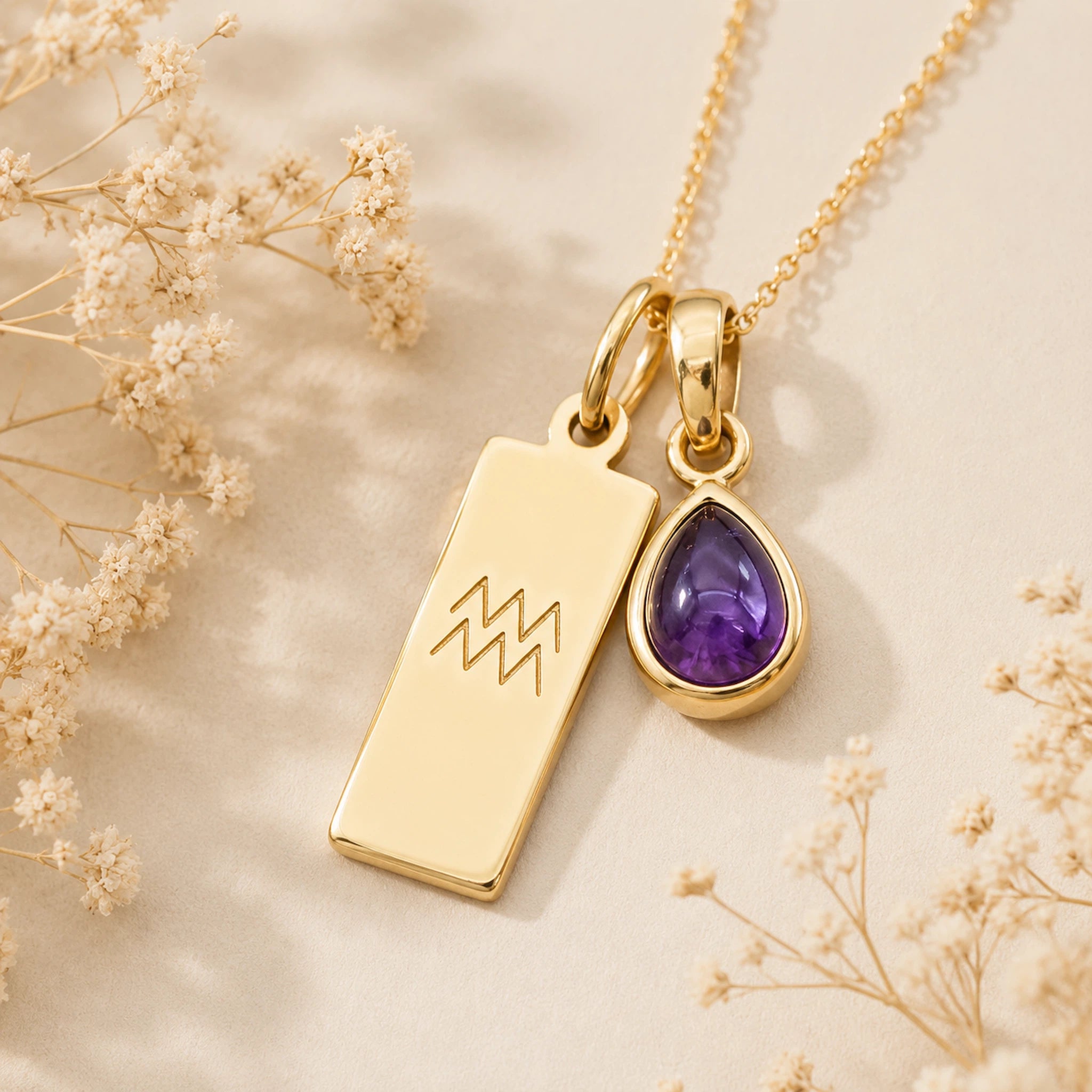 Gold necklace with a teardrop february birthstone natural amethyst pendant and engraved aquarius glyph tag on a beige background with dried flowers.