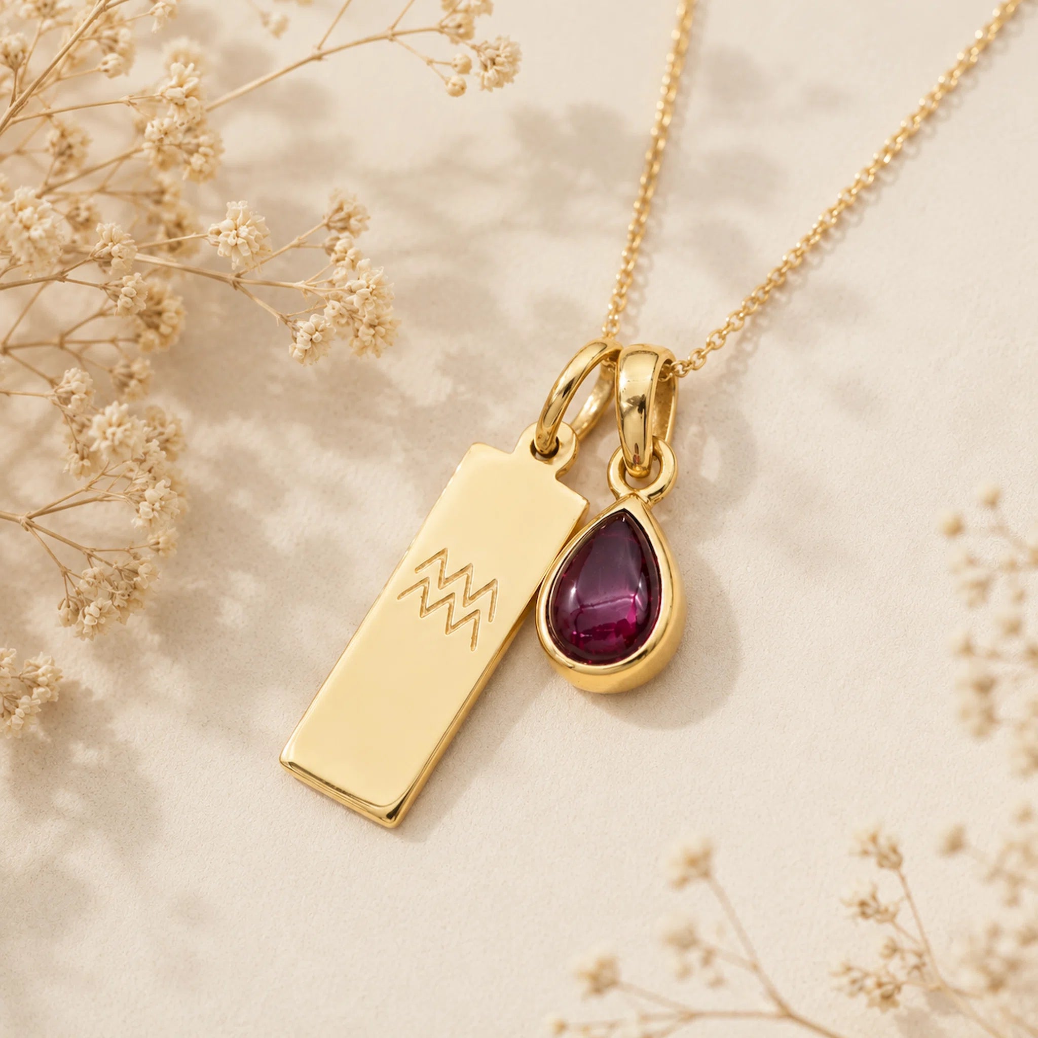Gold necklace with a teardrop january birthstone natural garnet pendant and engraved aquarius glyph tag on a beige background with dried flowers.
