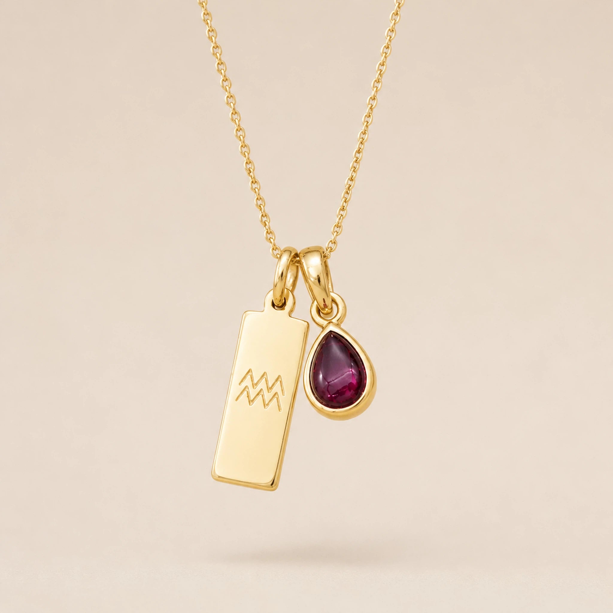 Gold necklace with a teardrop january birthstone natural garnet pendant and engraved aquarius glyph tag hanging on a beige background.