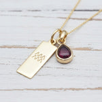Gold necklace with a teardrop-shaped garnet gemstone pendant and engraved aquarius zodiac symbol on a light wooden surface.