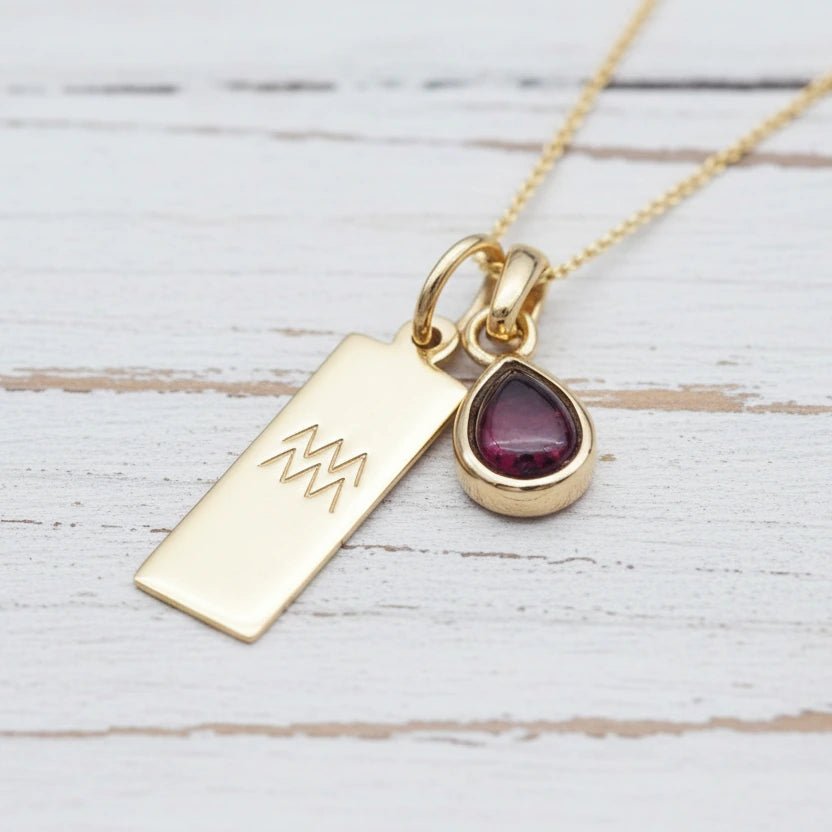 Gold necklace with a teardrop-shaped garnet gemstone pendant and engraved aquarius zodiac symbol on a light wooden surface.