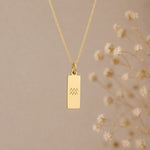 aquarius necklace - A gold rectangular pendant necklace with a simple engraved aquarius star sign line glyph symbol, hanging against a warm beige background 