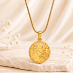 A gold aquarius zodiac sign pendant necklace, displayed on a textured stone slab with soft cream fabric in the background.