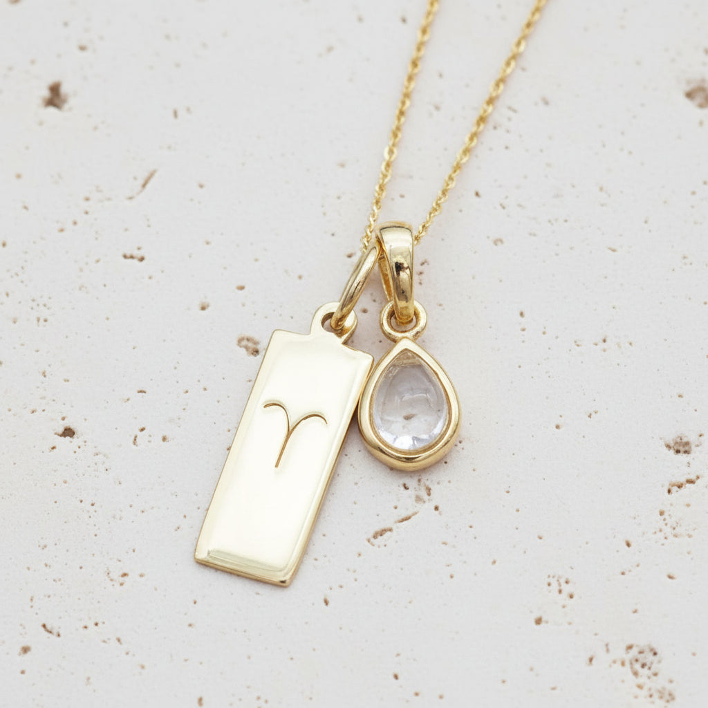 Gold necklace with Zodiac Aries pendant and april Birthstone herkimer quartz pendant on stone surface
