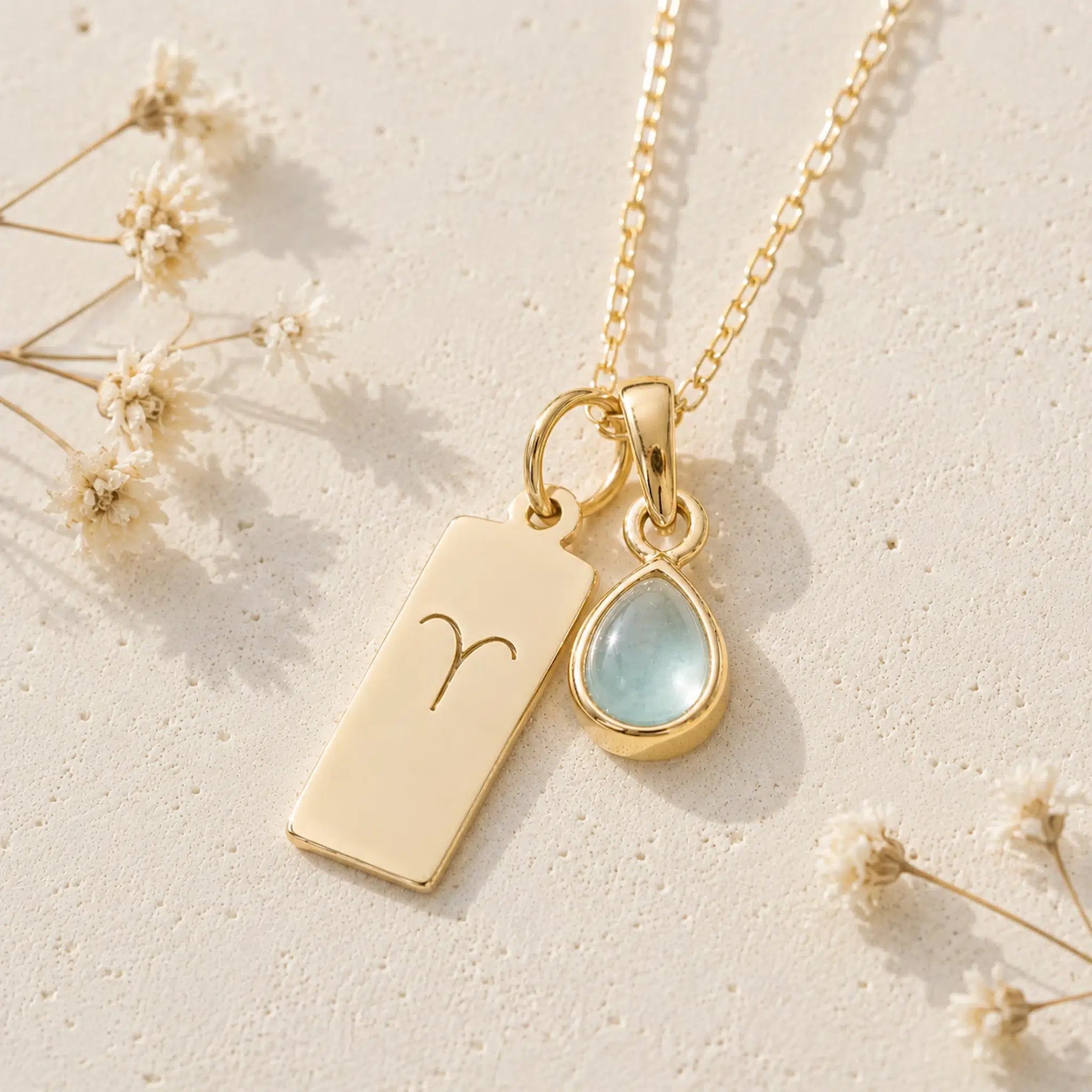 gold necklace with march birthstone aquamarine pendant and rectangle pendant with engraved aries line glyph on a light background.