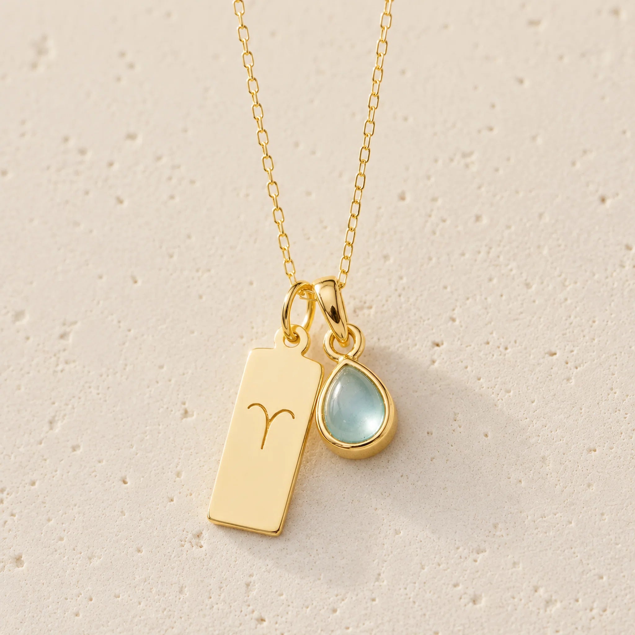 gold necklace with march birthstone aquamarine pendant and rectangle pendant with engraved aries line glyph hanging on a light background.