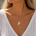 gold necklace with march birthstone aquamarine pendant and rectangle pendant with engraved aries line glyph worn by a person with a white shirt.