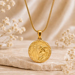 A gold pendant necklace with a circular aries zodiac sign design, resting on a textured stone with soft dried flowers in a warm, neutral setting.