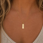 A close-up of a woman's neck and chest wearing a delicate gold chain necklace with a small rectangular pendant engraved with the aries zodiac symbol.