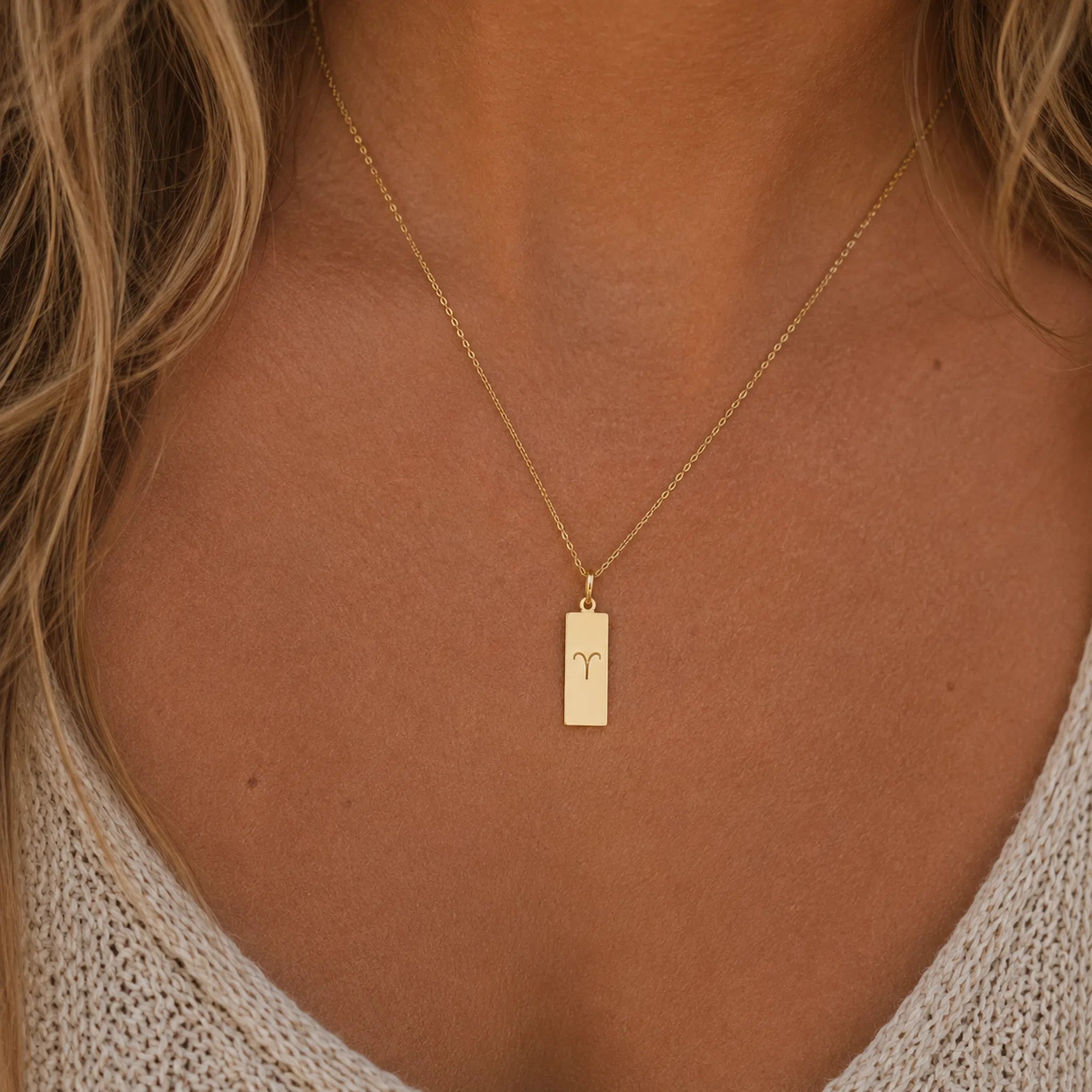 A close-up of a woman's neck and chest wearing a delicate gold chain necklace with a small rectangular pendant engraved with the aries zodiac symbol.