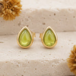 A pair of August Birthstone Natural Peridot Earrings - gold teardrop stud earrings with green gemstone centers, displayed on a textured beige stone surface with dried flowers.