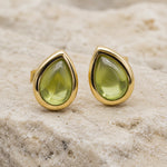 A pair of August Birthstone Natural Peridot Earrings - gold teardrop stud earrings with green gemstones, resting on a textured beige stone surface.