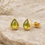 A pair of August Birthstone Natural Peridot Earrings - gold teardrop stud earrings with smooth olive-green cabochon gemstones, displayed on a textured light beige stone surface with dried flowers in the background.