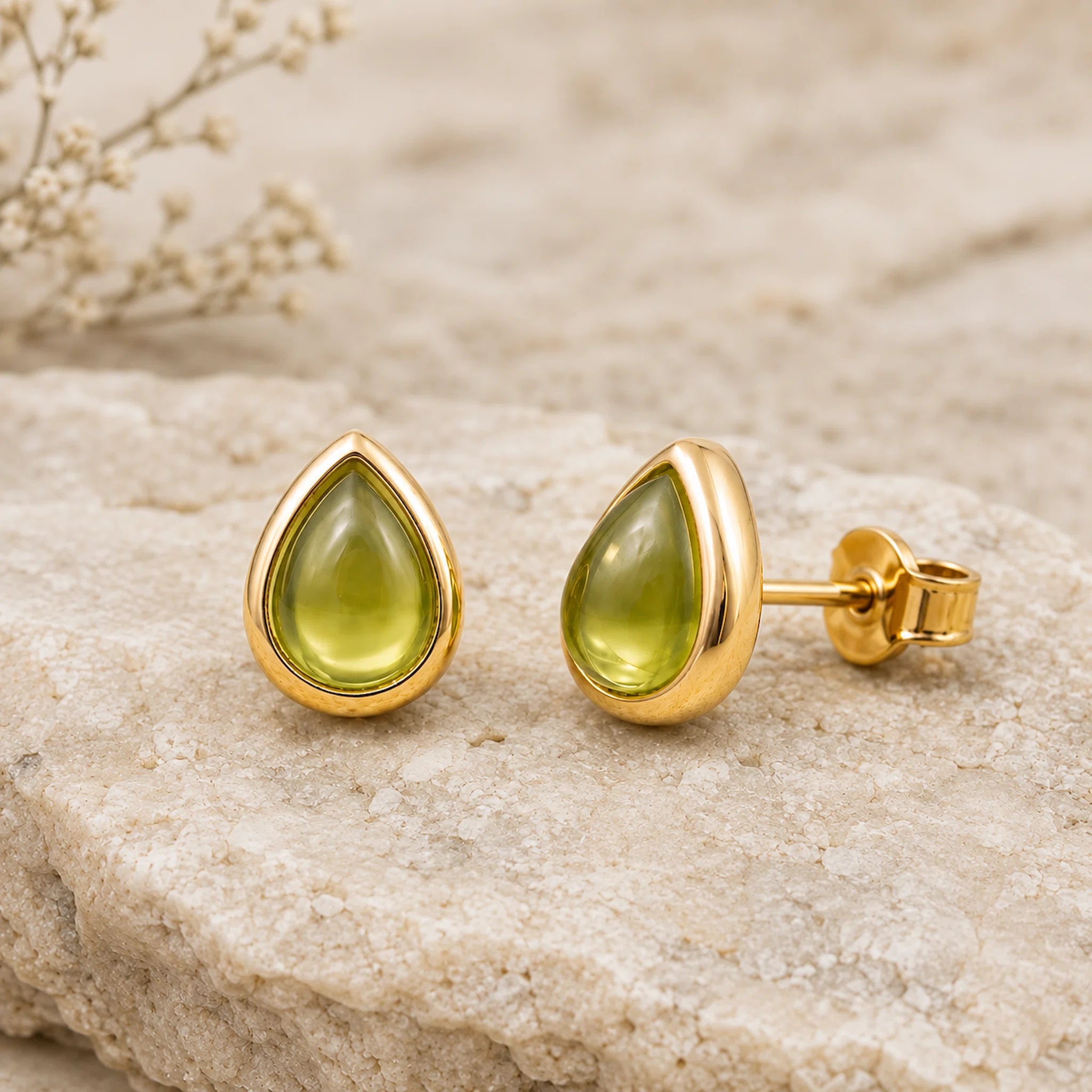 A pair of August Birthstone Natural Peridot Earrings - gold teardrop stud earrings with smooth olive-green cabochon gemstones, displayed on a textured light beige stone surface with dried flowers in the background.