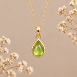 A gold august birthstone necklace featuring a teardrop pendant with a polished green natural peridot gemstone, suspended from a delicate gold chain against a warm beige background with soft white dried flowers.