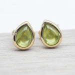 Gold stud earrings with green peridot teardrop stones on a light wooden surface