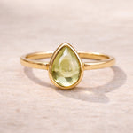 Gold august birthstone ring with a teardrop-shaped green natural peridot gemstone on a light background
