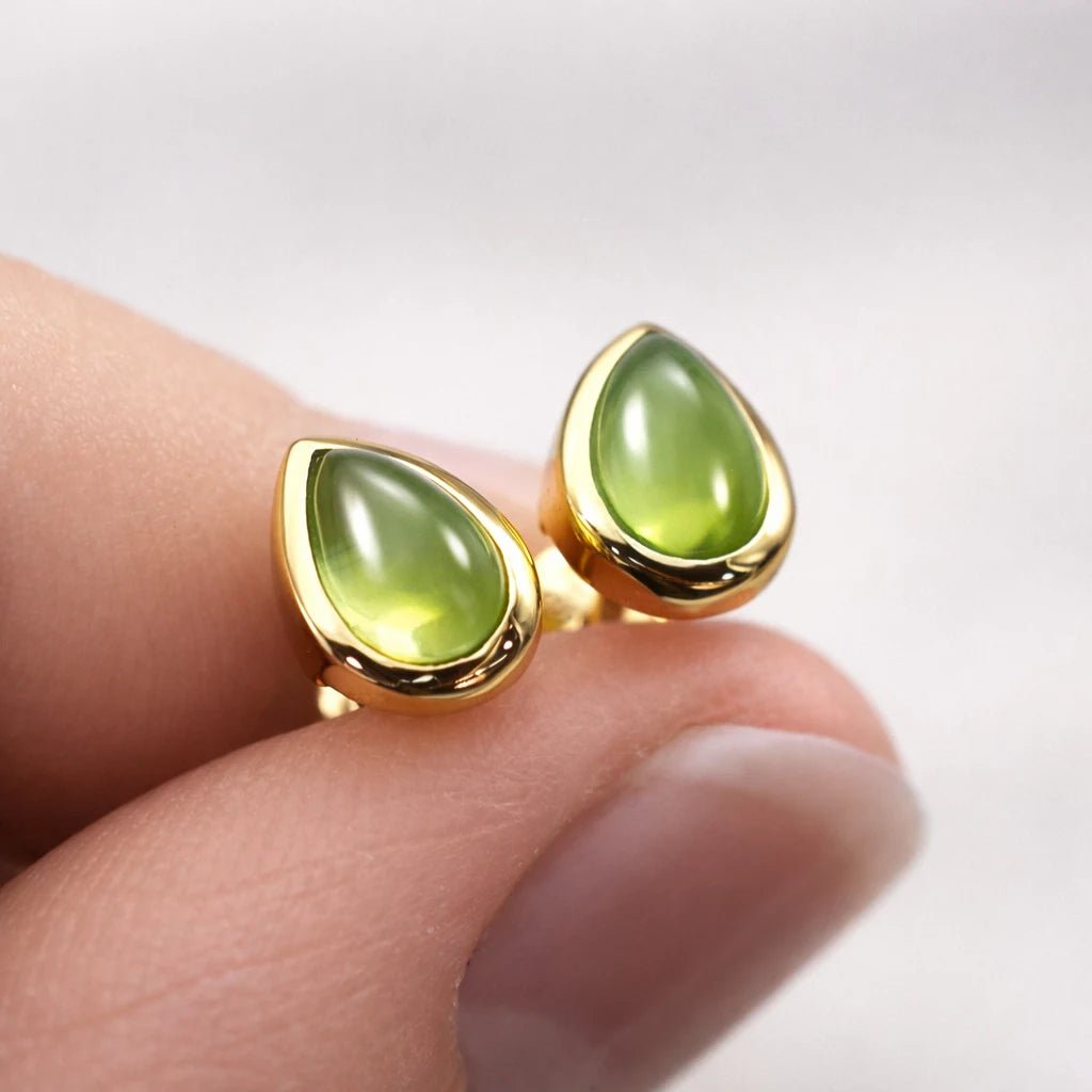 Gold stud earrings with green natural peridot gemstones held between fingers on a light background