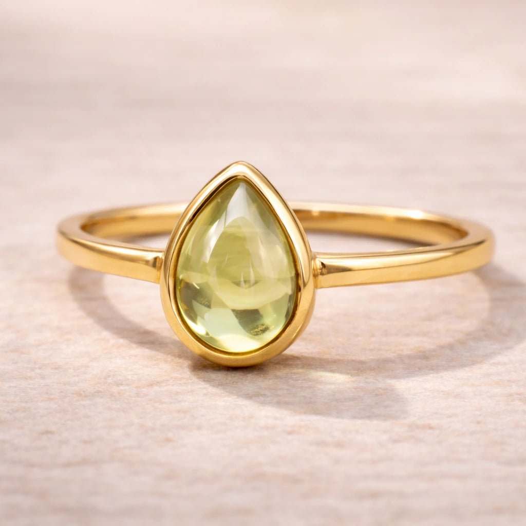 Gold plated August birthstone ring with a pear-shaped light green natural peridot gemstone cabochon in a polished low-profile bezel setting on a beige stone surface
