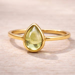 Gold plated August birthstone ring with a pear-shaped light green natural peridot gemstone cabochon in a polished low-profile bezel setting on a beige stone surface