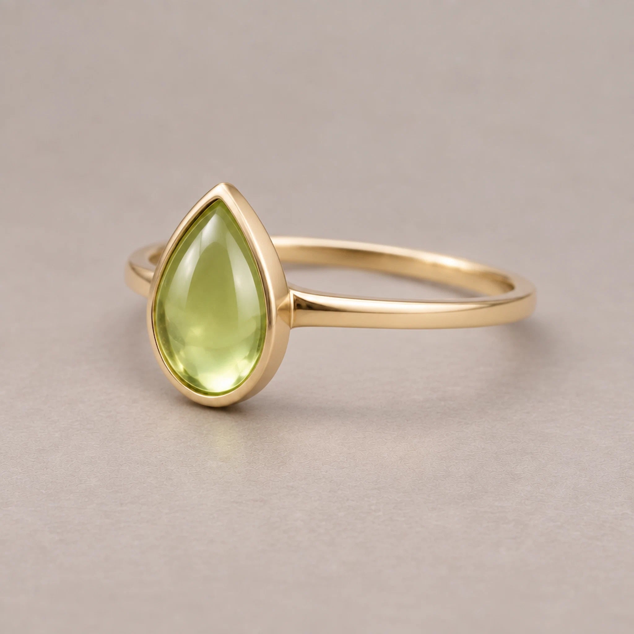 Gold plated August birthstone ring with a pear-shaped light green natural peridot gemstone cabochon in a polished low-profile bezel setting on a beige surface
