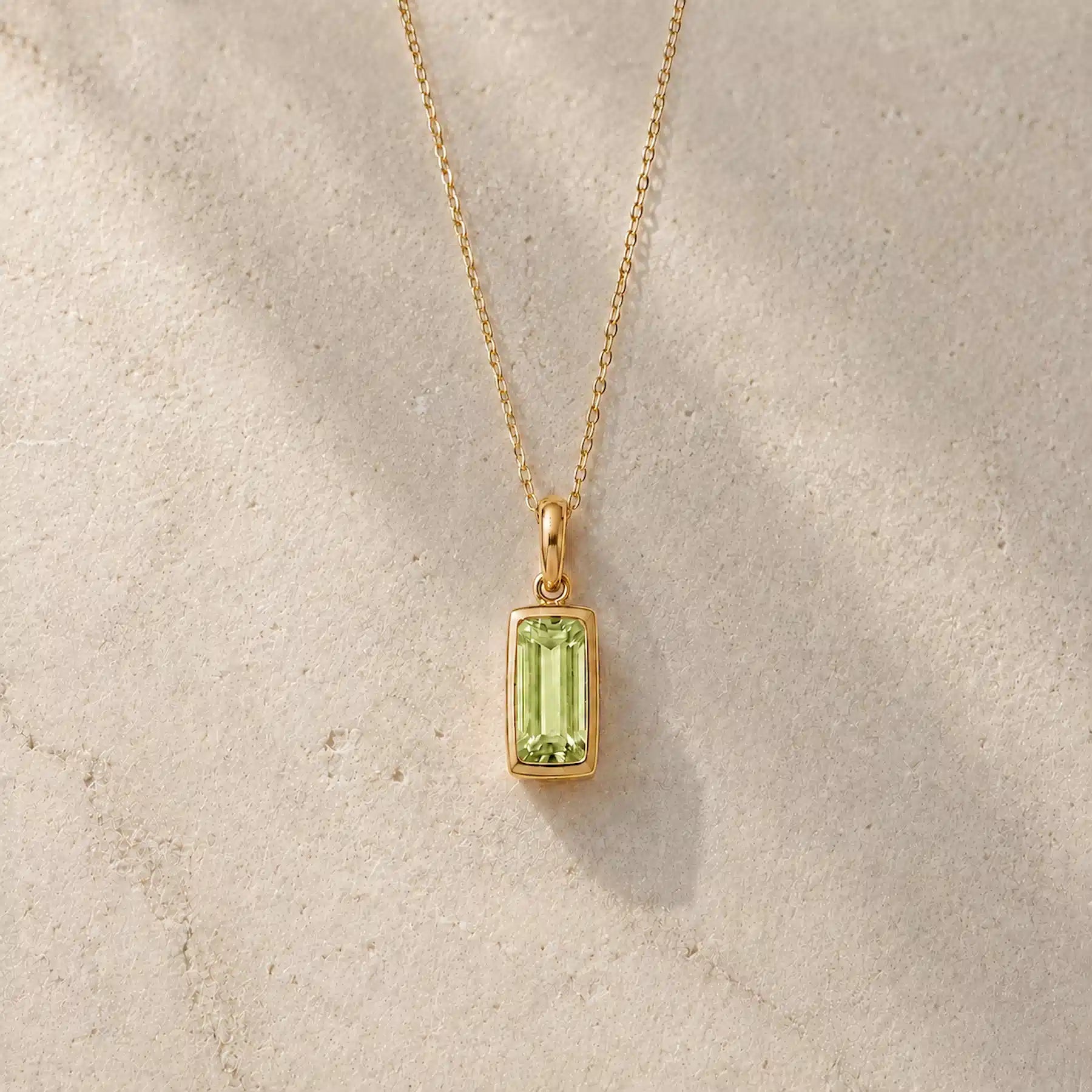 Gold fine chain necklace with a rectangle faceted bezel set august cubic zirconia pendant on a light stone background.