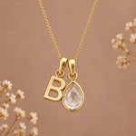 Gold initial pendant necklace featuring a polished gold letter 'B' and a teardrop-shaped clear crystal pendant, displayed against a warm brown background with delicate dried flowers.