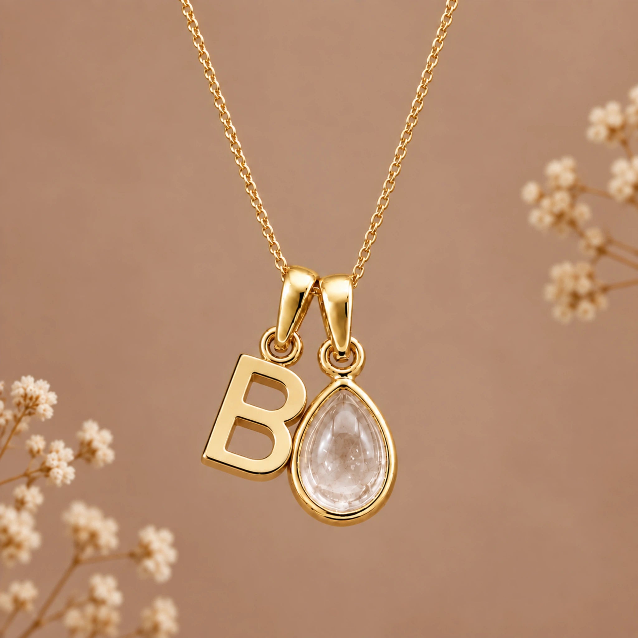 Gold initial pendant necklace featuring a polished gold letter 'B' and a teardrop-shaped clear crystal pendant, displayed against a warm brown background with delicate dried flowers.