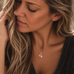 A close-up lifestyle shot of a woman with blonde hair wearing a delicate gold chain necklace with a gold initial 'B' pendant and a small teardrop crystal charm.