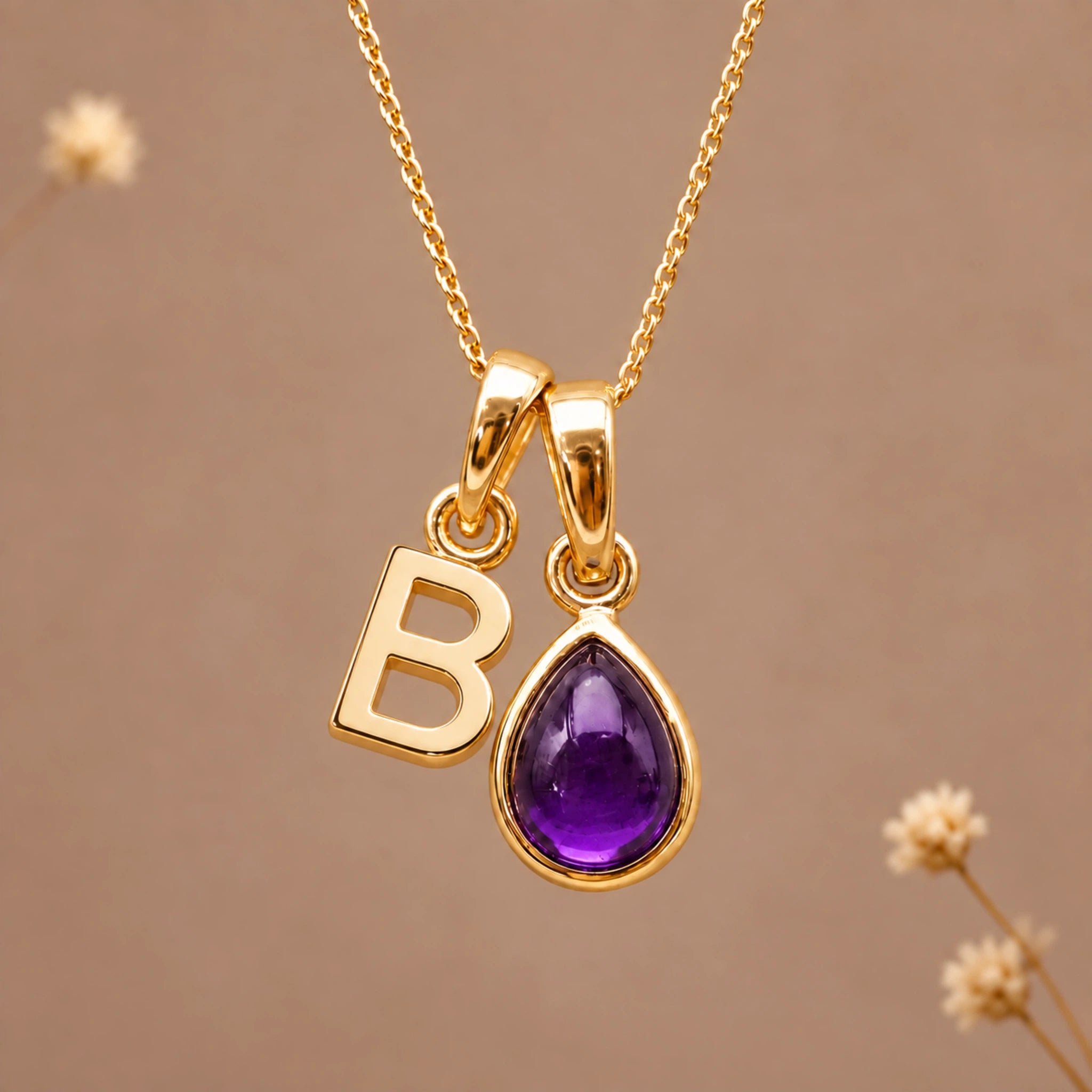 Gold initial pendant necklace featuring the letter 'B' and a february birthstone teardrop-shaped natural amethyst gemstone charm, displayed against a warm beige background with subtle dried flowers.