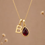 Gold initial pendant necklace featuring the letter 'B' and a teardrop-shaped dark red january birthstone natural garnet gemstone charm, displayed against a warm beige background with subtle dried flowers.