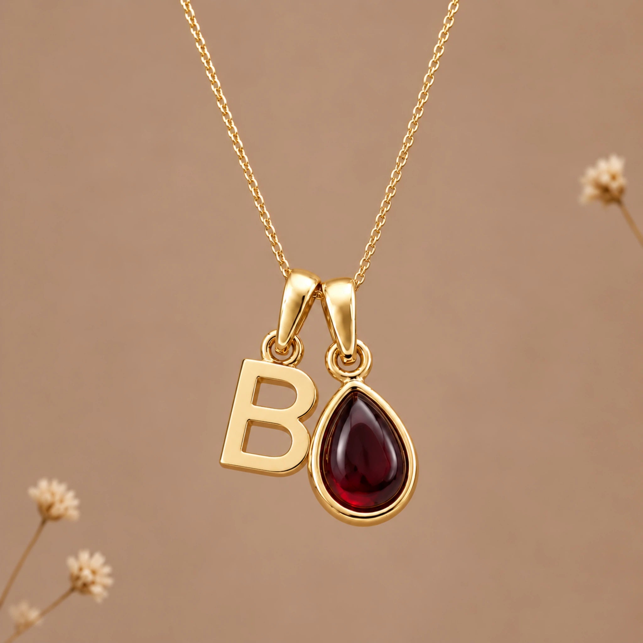 Gold initial pendant necklace featuring the letter 'B' and a teardrop-shaped dark red january birthstone natural garnet gemstone charm, displayed against a warm beige background with subtle dried flowers.
