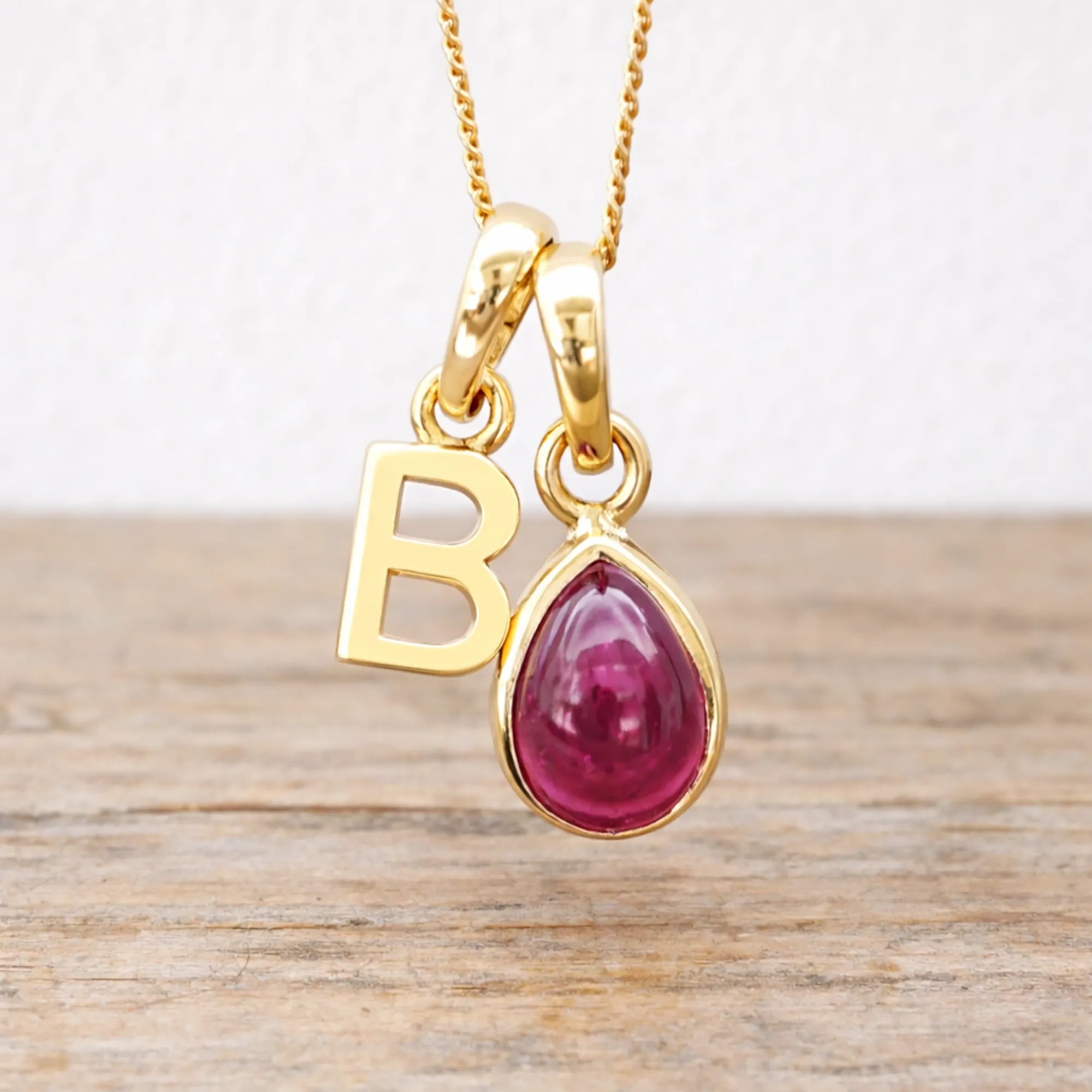 Gold necklace with a red ruby gemstone pendant and 'B' letter charm on a wooden surface.