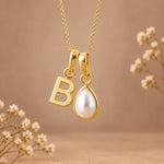 A gold initial pendant necklace featuring a letter 'B' and a teardrop-shaped white pearl, shown against a warm beige background with delicate dried flowers.