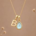 A gold initial march birthstone necklace featuring a letter 'B' pendant and a teardrop-shaped light blue natural aquamarine gemstone pendant on a delicate gold chain, set against a warm beige background with subtle dried flowers.