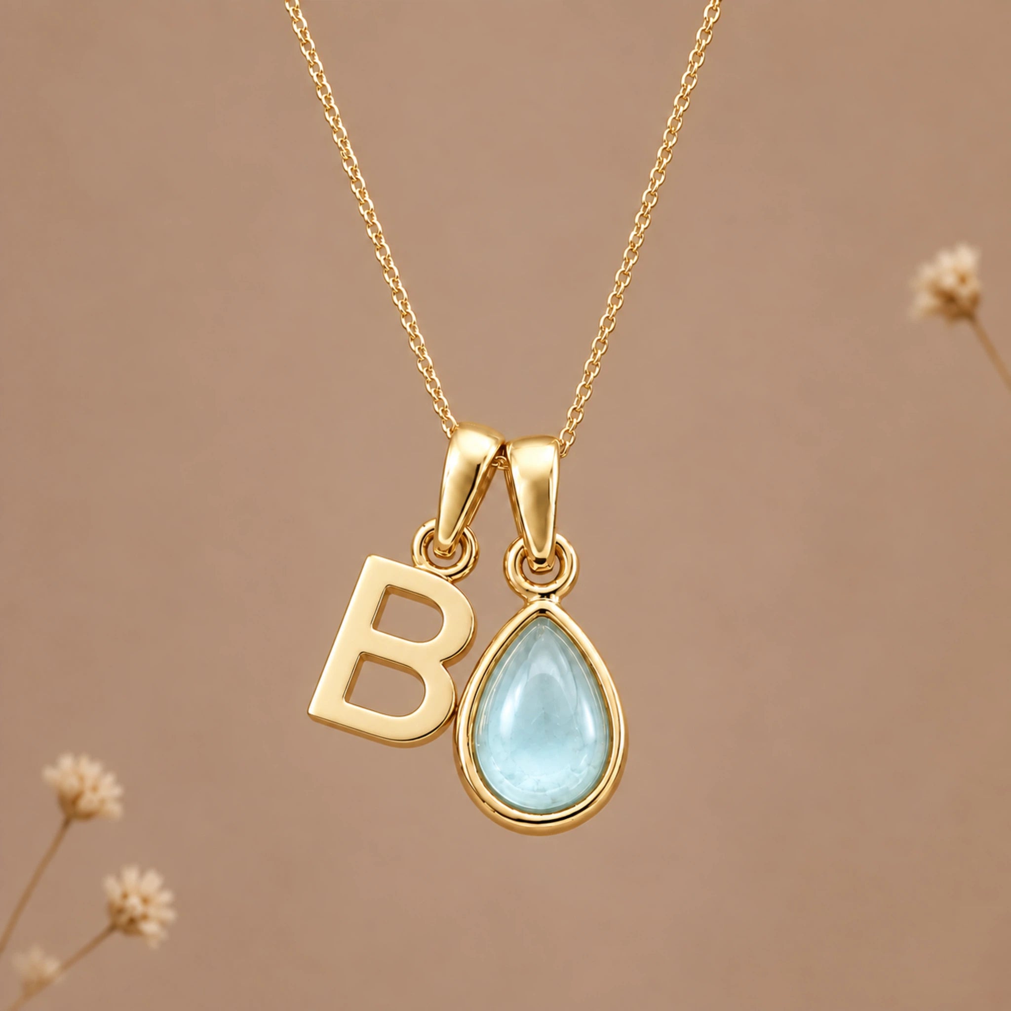 A gold initial march birthstone necklace featuring a letter 'B' pendant and a teardrop-shaped light blue natural aquamarine gemstone pendant on a delicate gold chain, set against a warm beige background with subtle dried flowers.