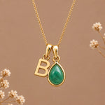Gold initial necklace with a teardrop-shaped green stone pendant and a gold letter 'B' charm, displayed on a warm brown background with dried flowers.